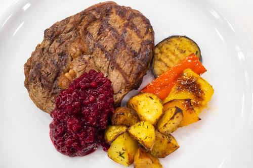 На фотографии представлен изысканный стейк из мяса, аккуратно размещенный на белой тарелке, великолепно дополненный свежими овощами и аппетитным брусничным соусом. Это блюдо олицетворяет высокое кулинарное искусство и внимание к деталям. Идеальное изображение для ресторанов, кулинарных мастер-классов и рекламы продуктов питания. Отражает великолепие гастрономического опыта.